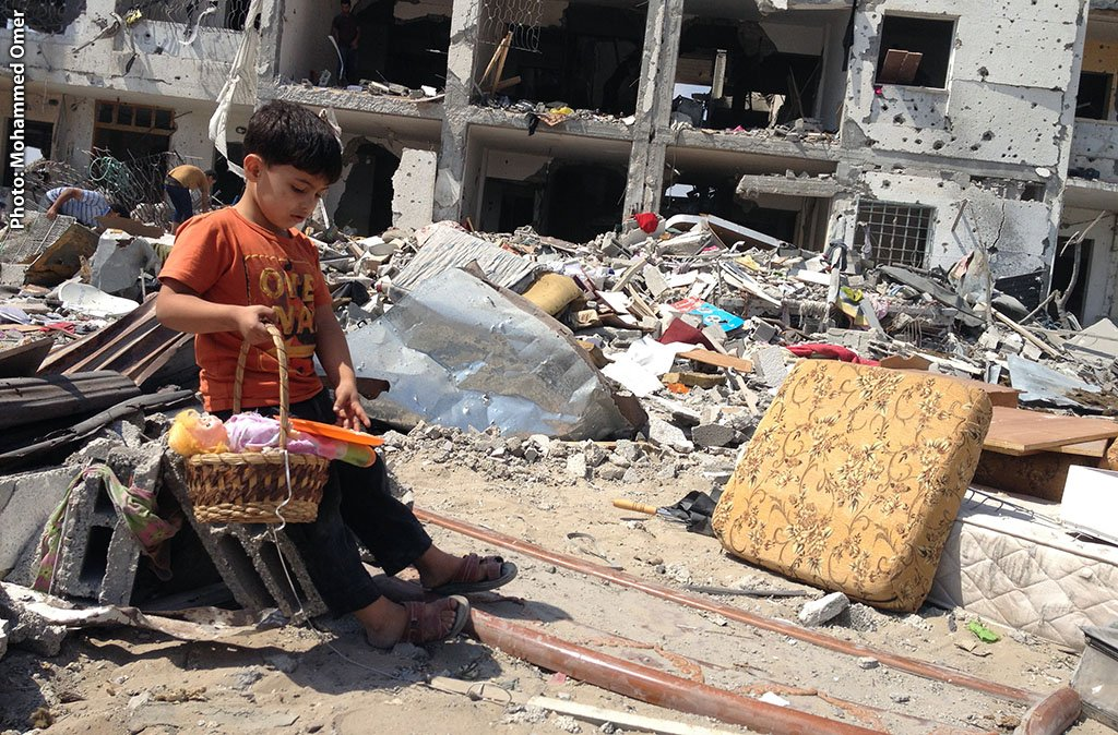 A young boy wearing a yellow orange T-shirt carrying a Basket of toys He's sitting next to the rubble on a bomb out residential building

 Photo by Mohamed Omer