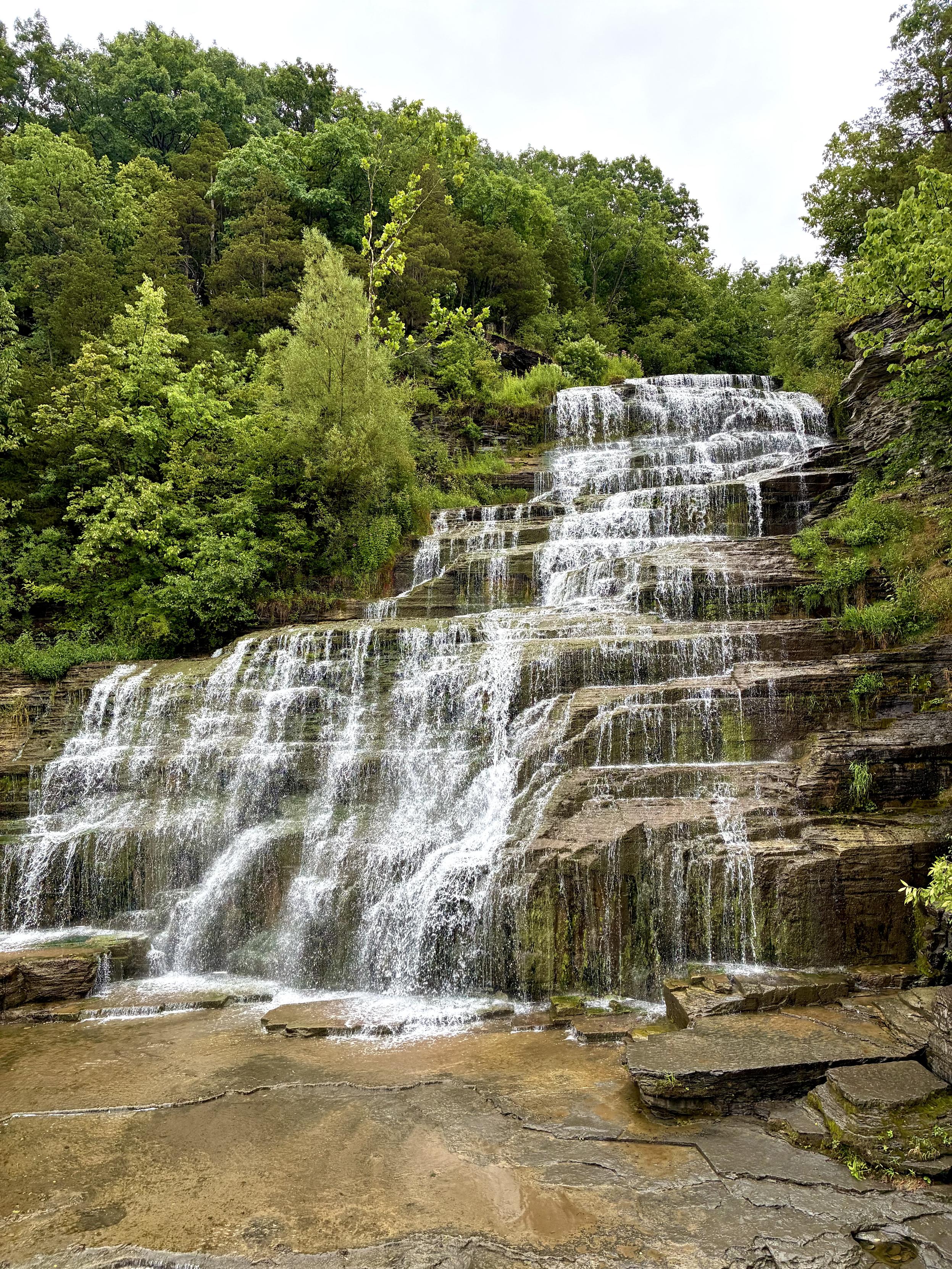 A broad, multi-tiered waterfall cascades over layers of flat rock, surrounded by dense green forest. Water fans across the wide rocky ledges before flowing onto a shallow stone bed at the base.