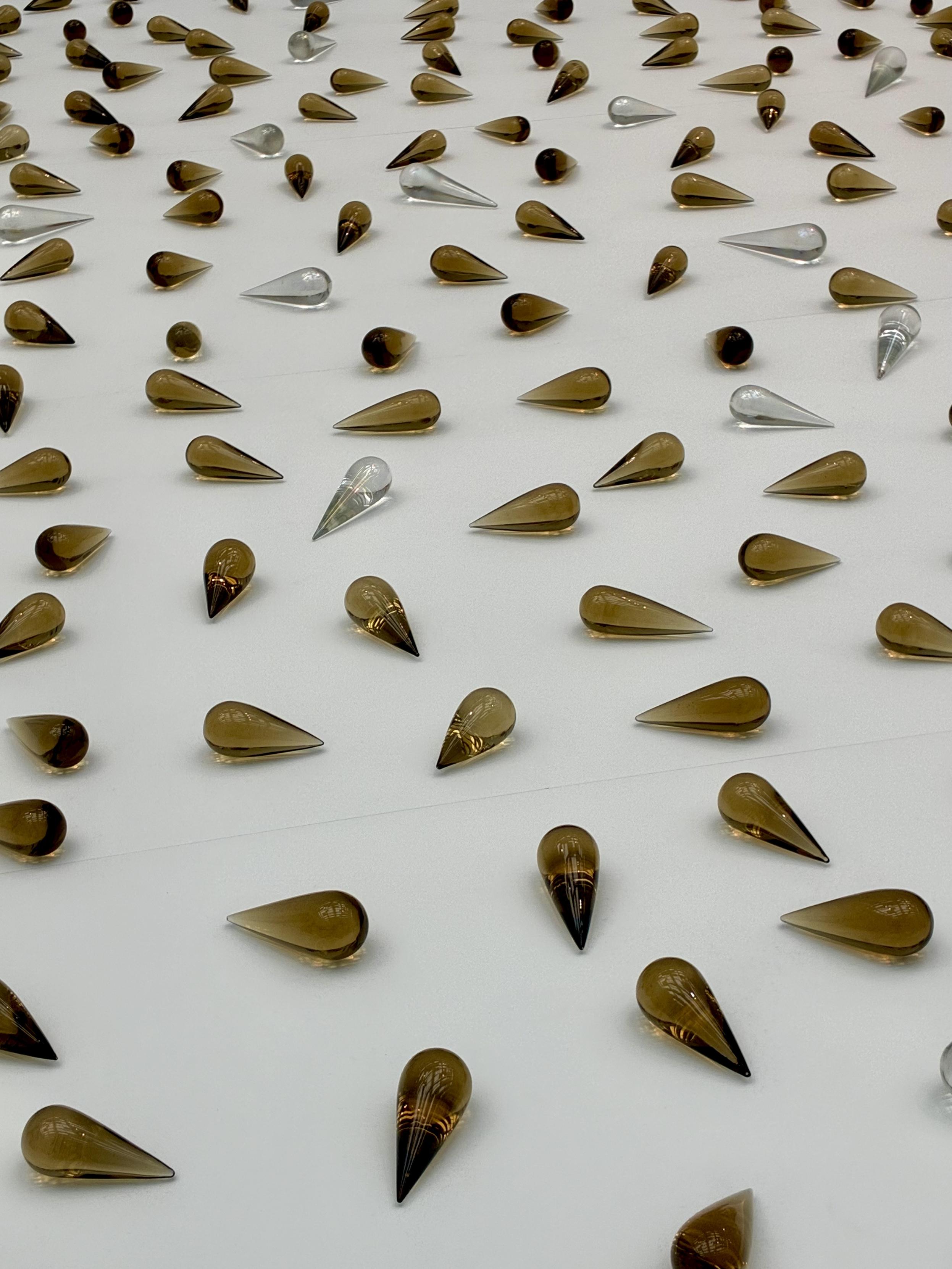 Dozens of amber and clear teardrop-shaped glass pieces spread across a white surface, forming an abstract representation of a flowing stream.