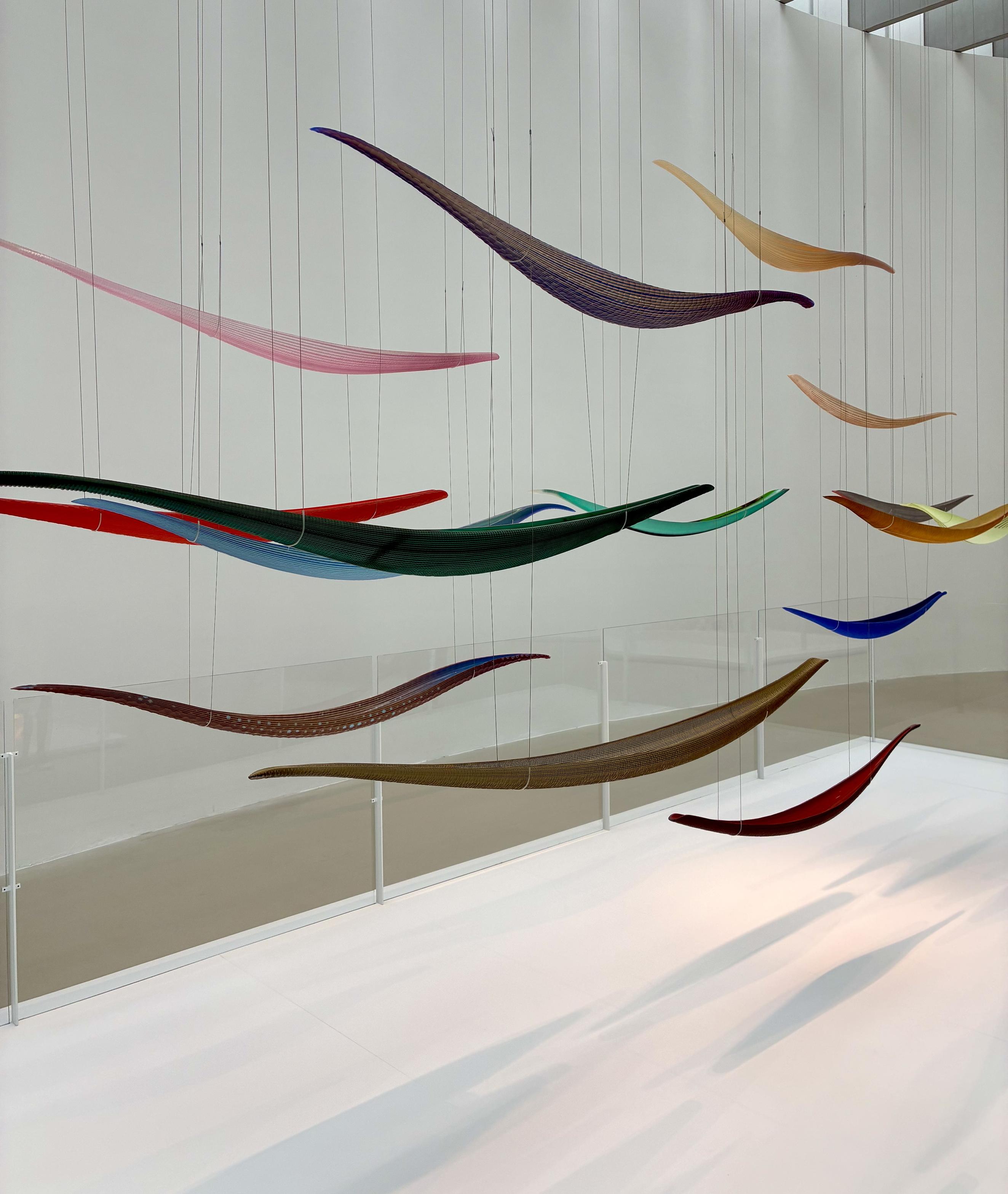 Colorful, elongated glass sculptures hang from thin wires in a bright, modern gallery, appearing to float through the air like a school of fish.