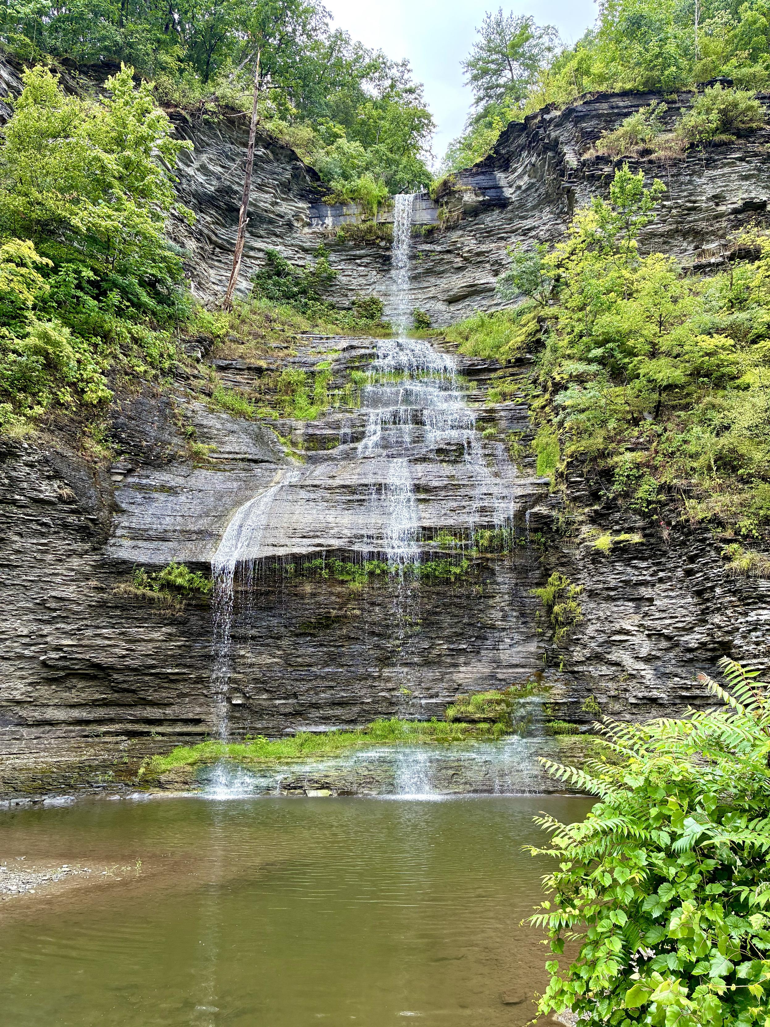 A tall, multi-tiered waterfall cascades over layered gray rock cliffs into a calm pool below, surrounded by lush green trees and vegetation.