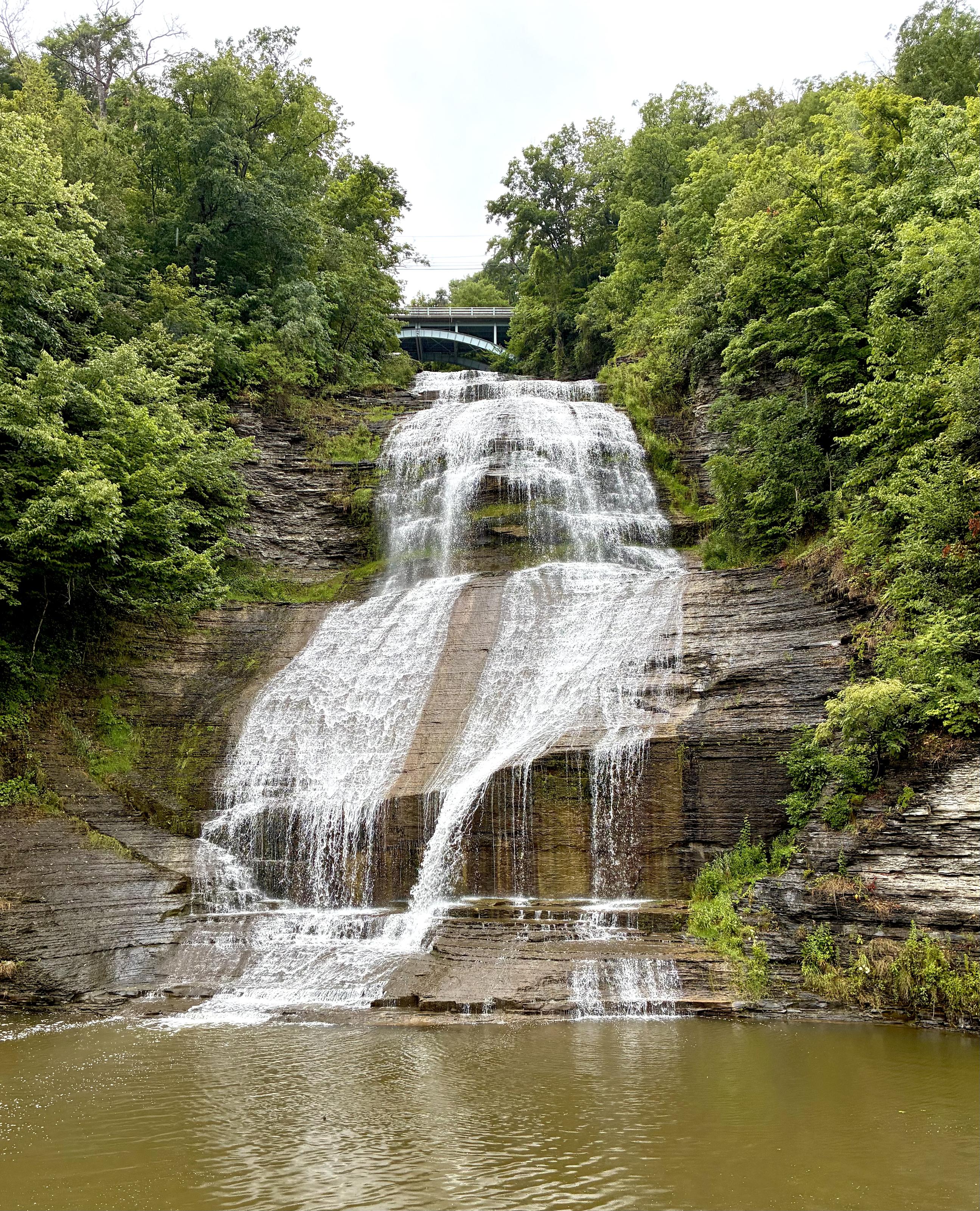 A broad, multi-tiered waterfall flows over smooth, layered rock into a large pool, framed by dense green forest, with a bridge visible above the falls.
