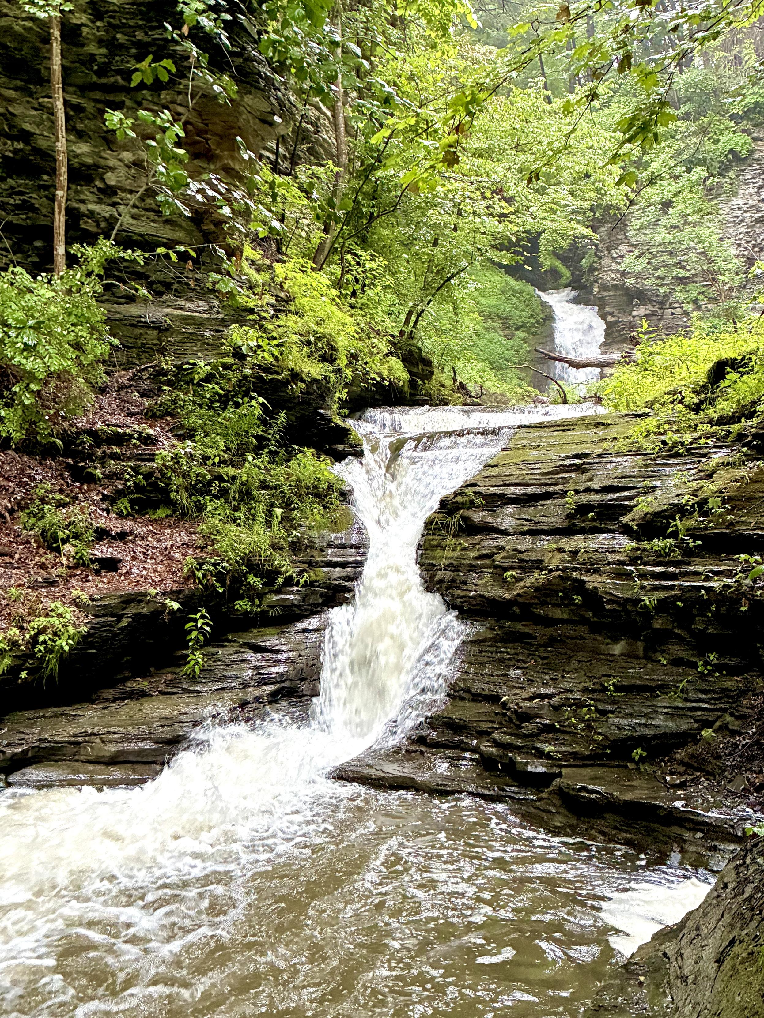 A rushing waterfall cascades over layered rock ledges through a lush green forest, with another waterfall visible farther upstream between the trees.