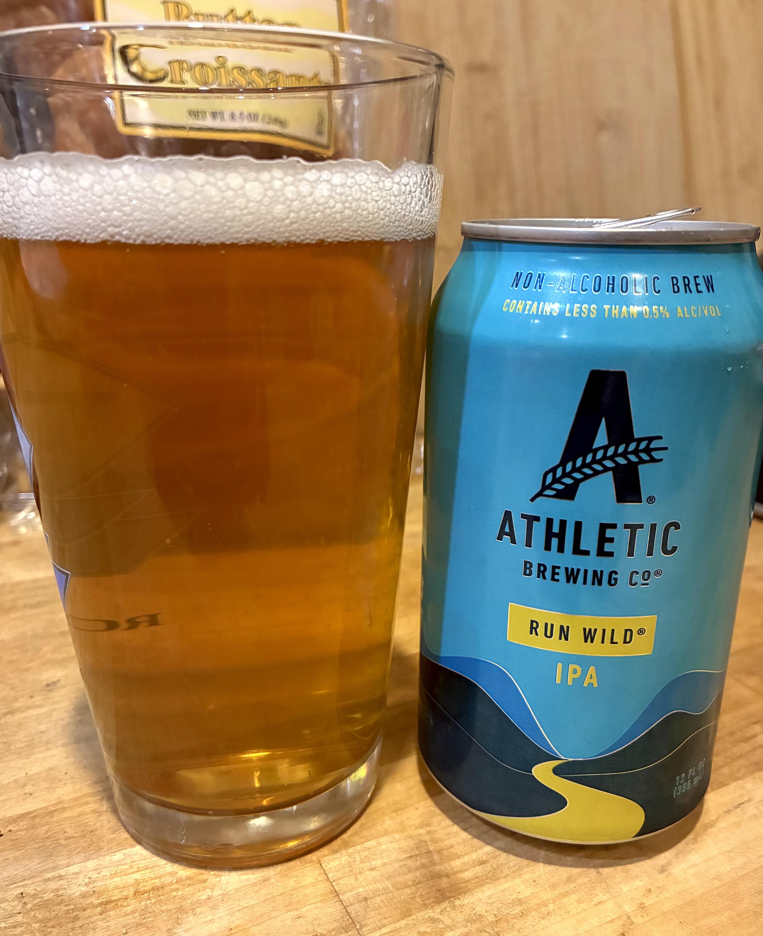 A pint glass filled with amber-colored beer with a foamy head sits next to a blue can of Athletic Brewing Co. Run Wild non-alcoholic IPA on a wooden table.