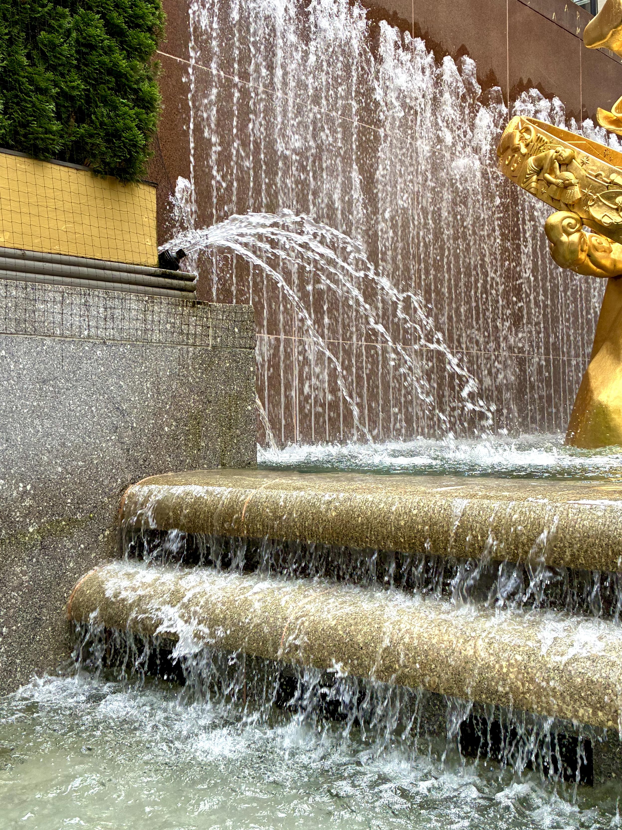 Water cascades over tiered stone ledges and sprays from fountains beside a large golden sculpture, creating a waterfall-like scene in the middle of the city.