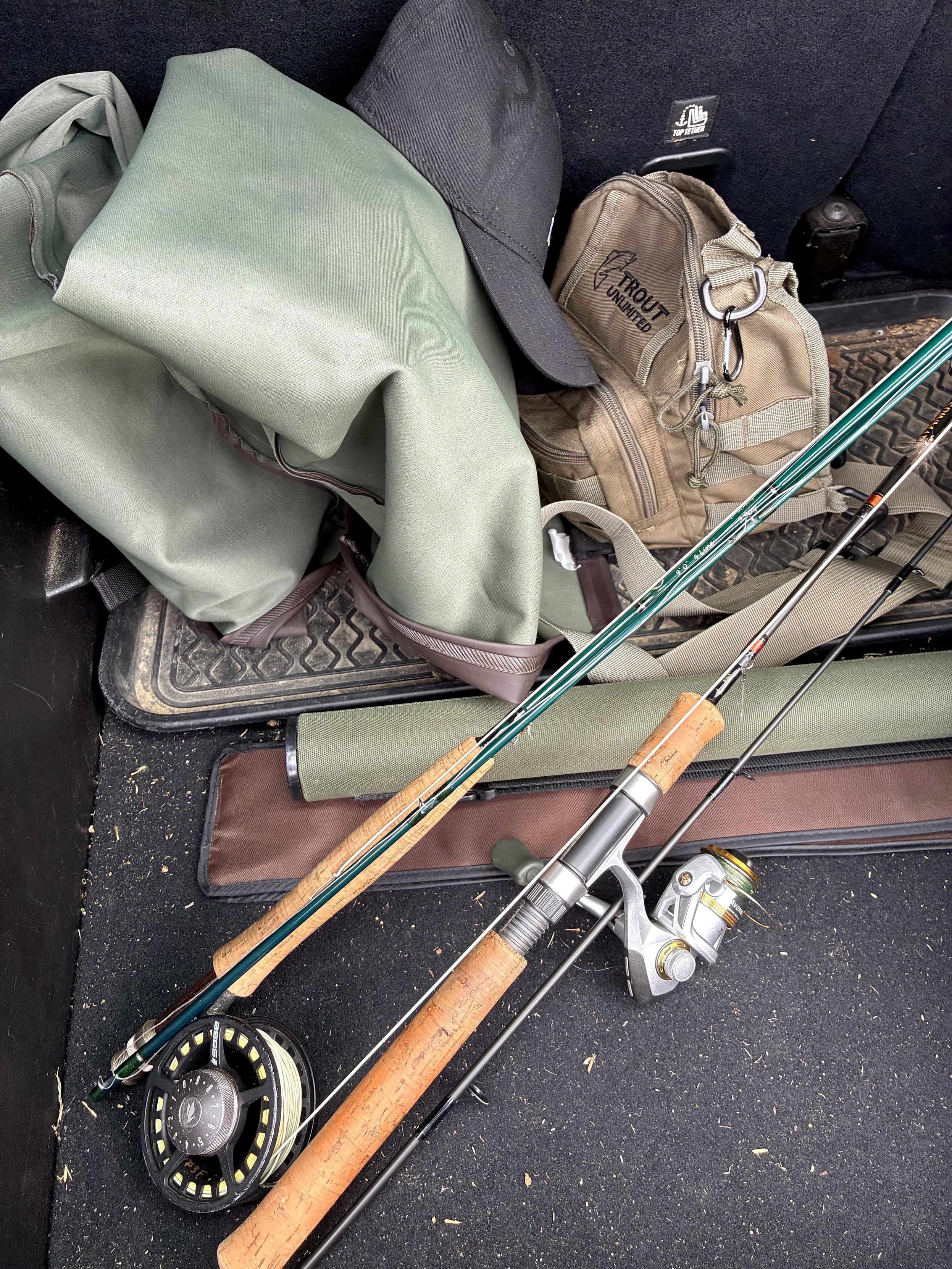 Two fishing rods, one with a spinning reel and one with a fly reel, lying in the trunk of a car alongside waders, a fishing bag, hat, and rod cases.