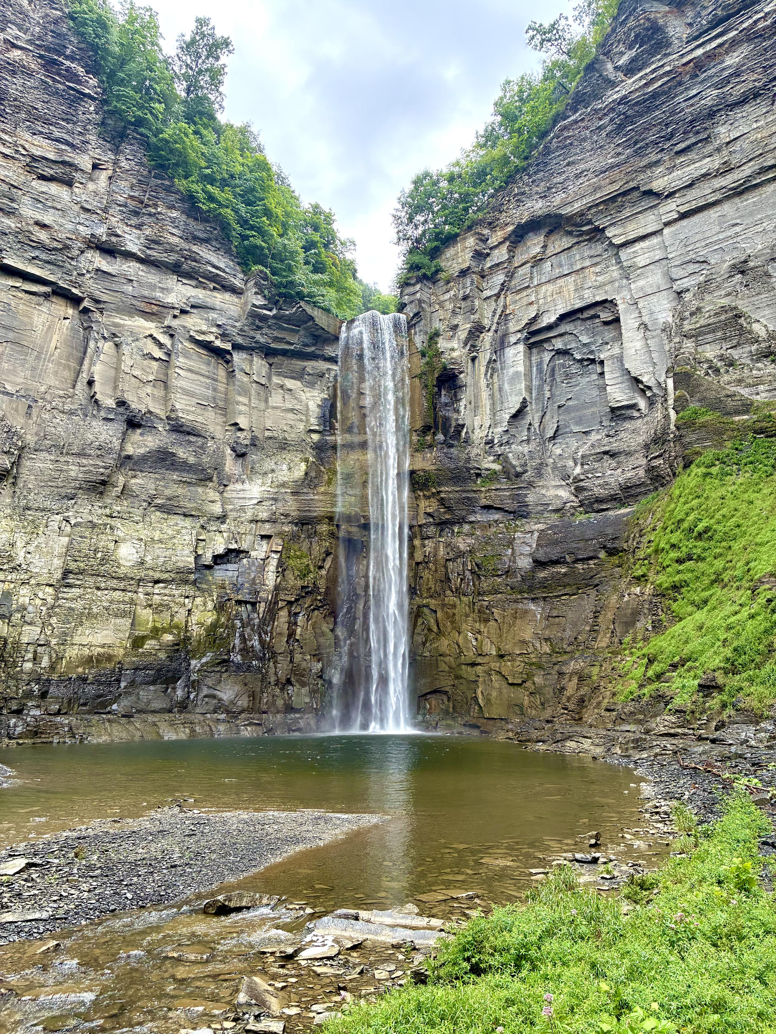 A tall waterfall plunging from a dramatic layered rock gorge into a broad pool below, surrounded by steep cliffs and lush green vegetation.